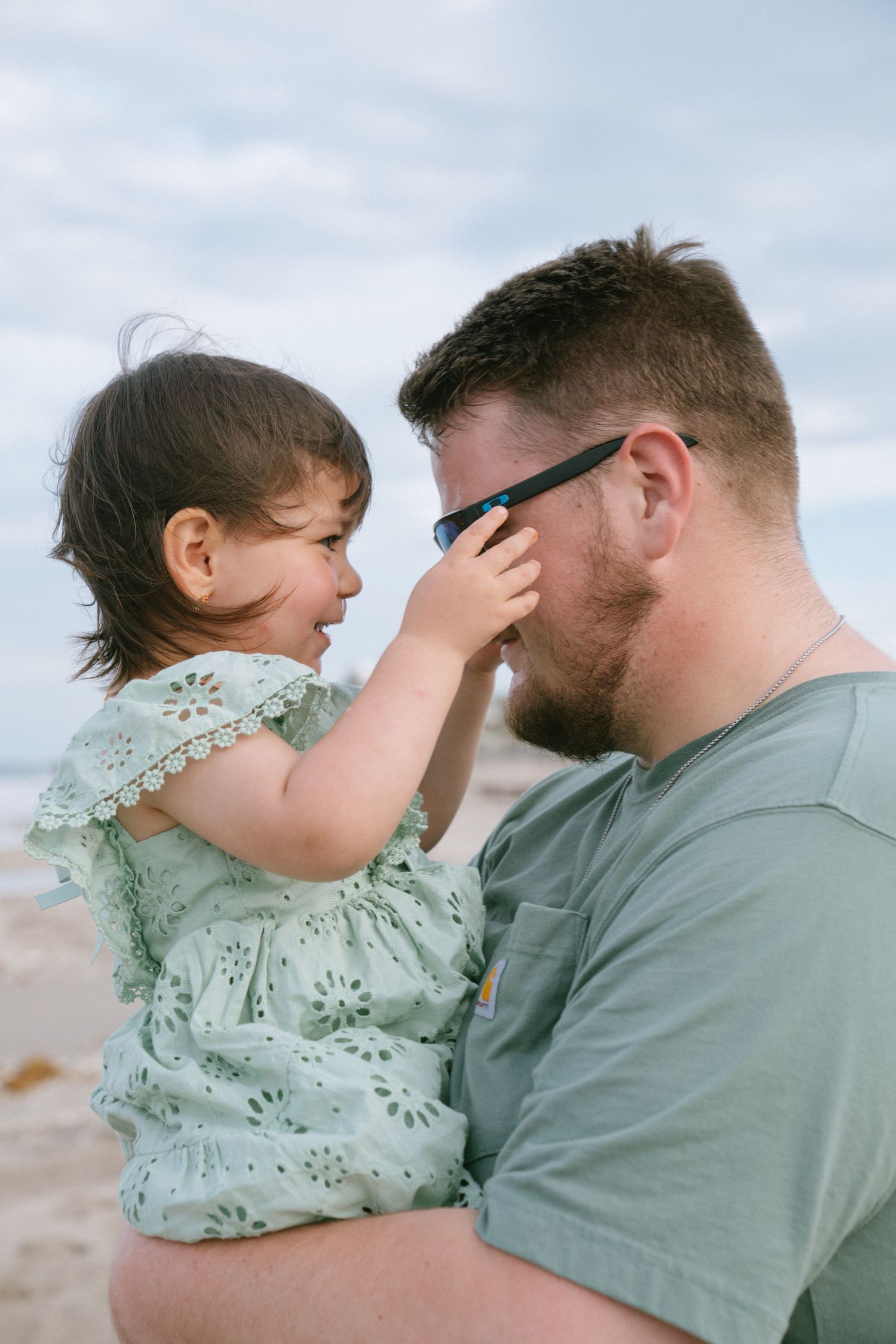 A bearded father holds his laughing toddler daughter on a beach, as she playfully touches his sunglasses.