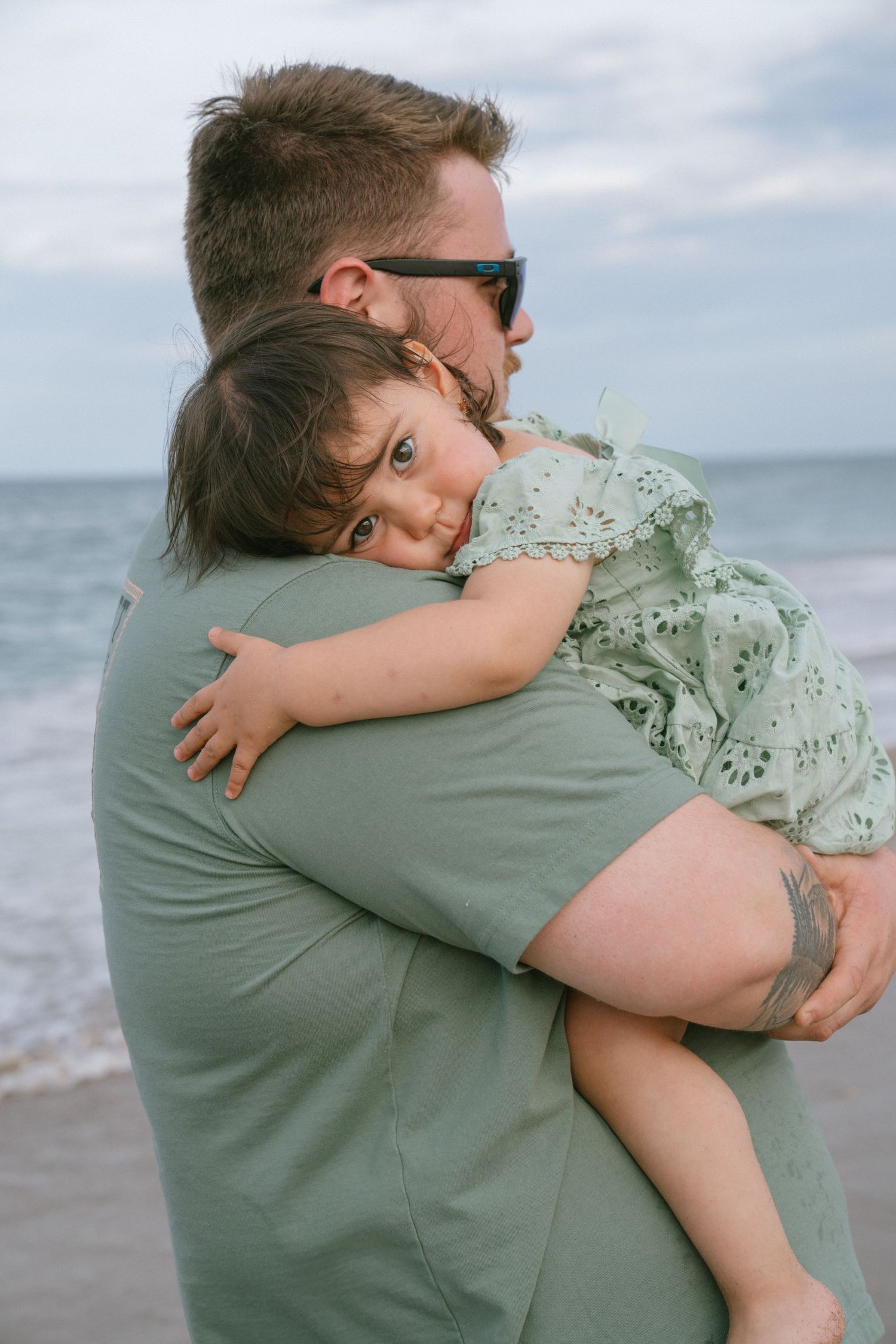 A father holds his young daughter on a beach, she rests her head on his shoulder in a tender embrace.