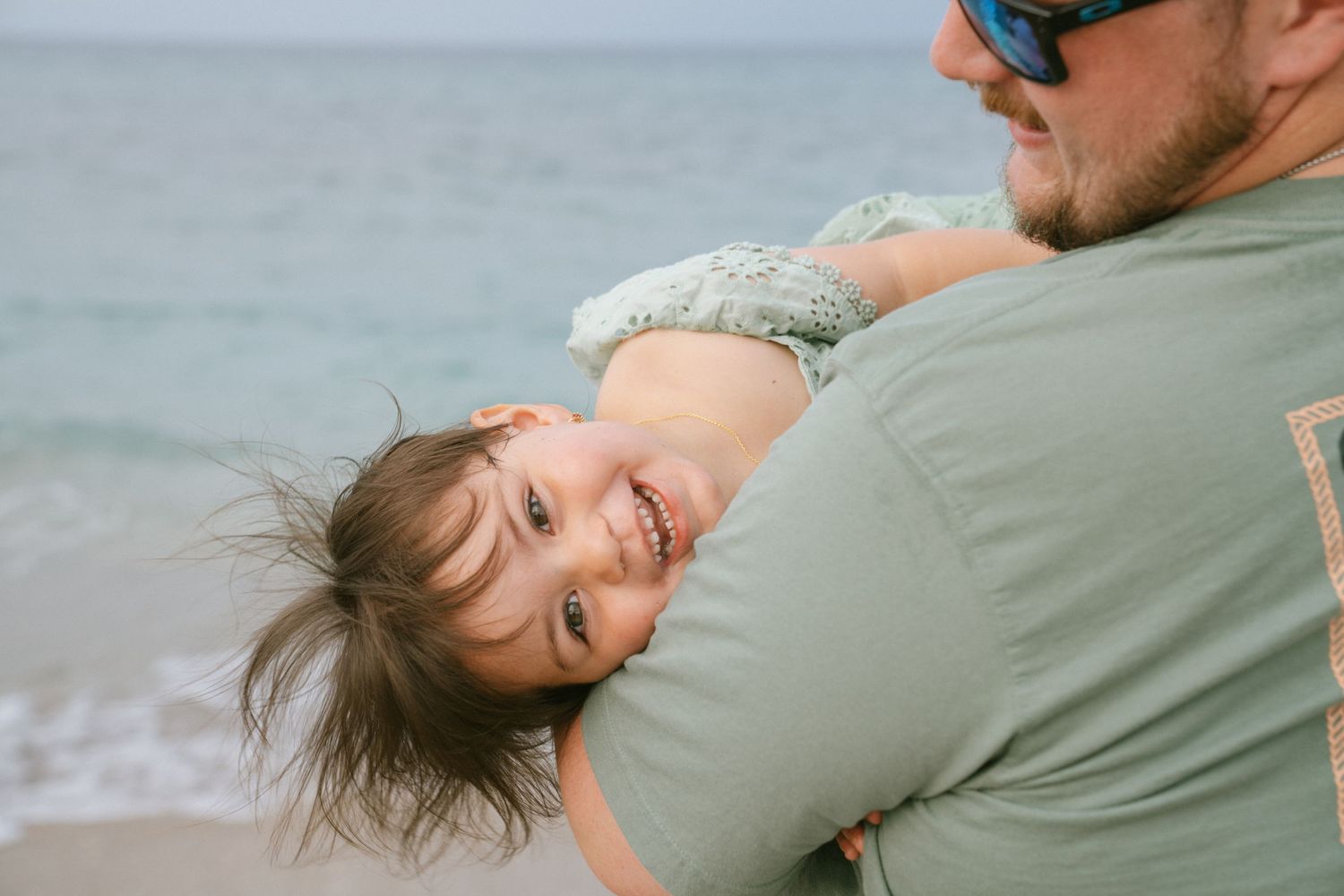 A joyful toddler laughs while being held by a man on a sandy beach with ocean waves in the background.