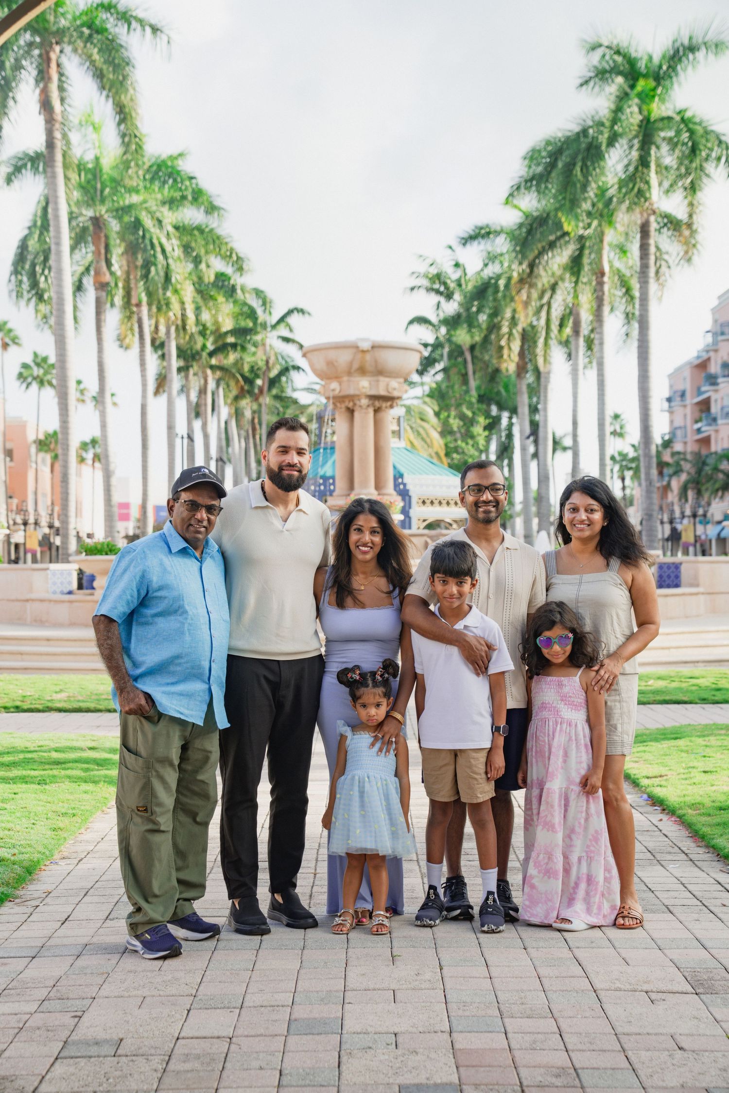 Multi-generational South Asian family posing together outdoors near a fountain lined with tall palm trees.