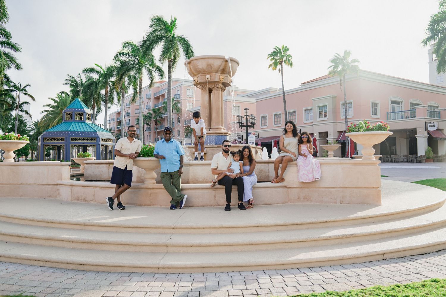 Family gathering around an ornate fountain in a tropical plaza with palm trees and pastel buildings in the background.
