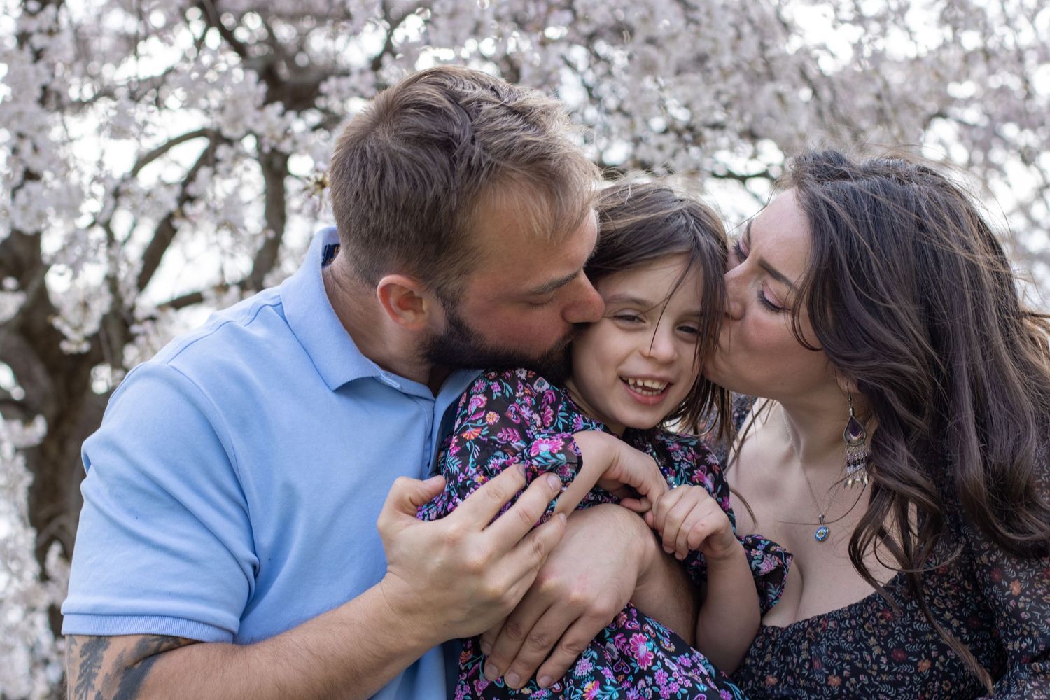 Family photographer in Cincinnati: Mom and dad kissing daughter during cherry blossom mini session at Ault Park. Outdoor portrait photograph