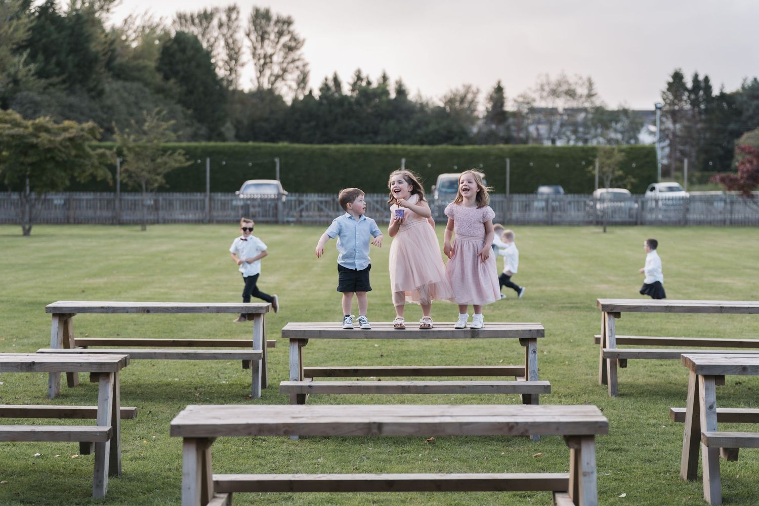 Children playing and standing on wooden picnic tables on a grassy field at sunset with trees in the background.