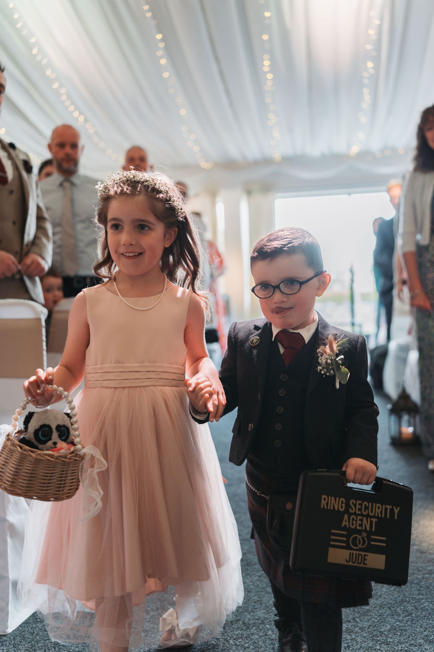 A young flower girl in pink dress and ring bearer in black suit walk down the aisle at a wedding ceremony under fairy lights.