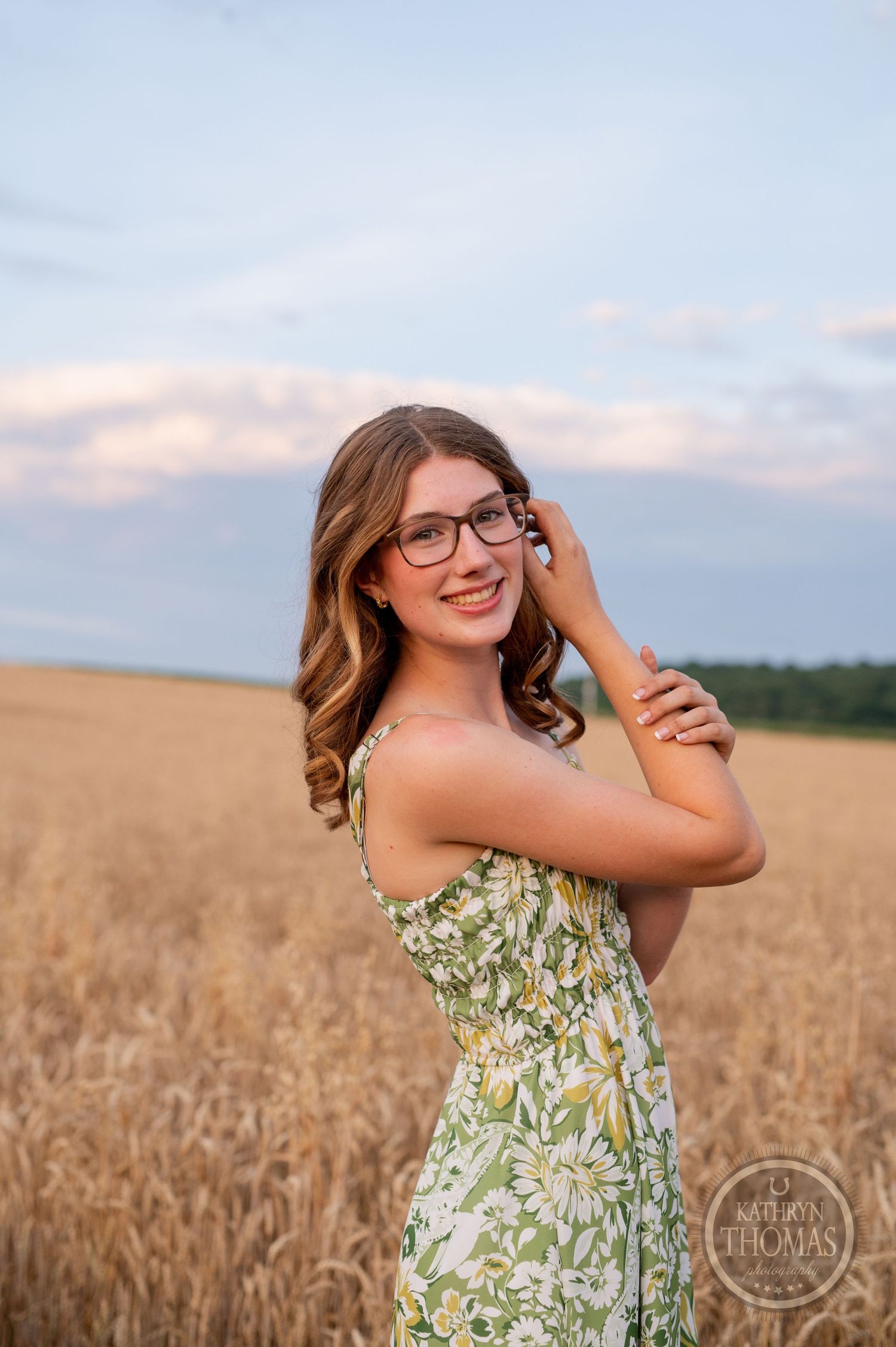 Portrait of someone in floral dress and glasses smiling in wheat field during golden hour.