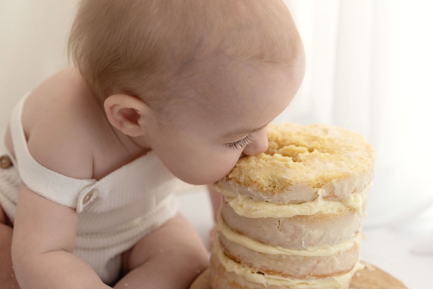 Baby in white clothing gazes curiously at a tall layered vanilla cake with cream filling.