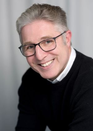 Professional headshot of a person wearing glasses, a black sweater and white collar, smiling warmly against a light background.