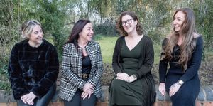 A group of four people sit together outdoors while smiling and laughing on a wooded bench.