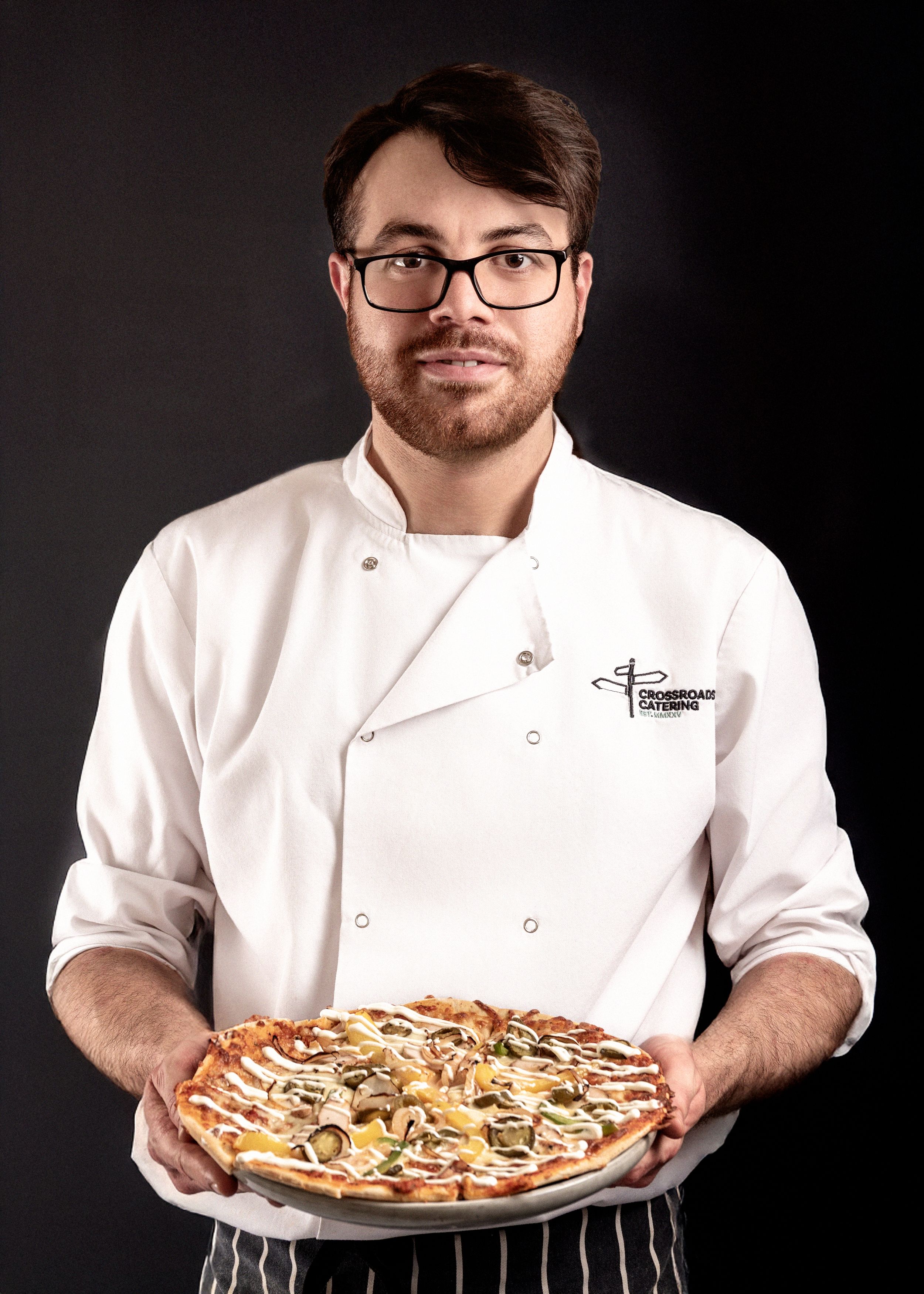 Professional chef in white coat displays a freshly made pizza topped with mushrooms on a round plate against dark background.