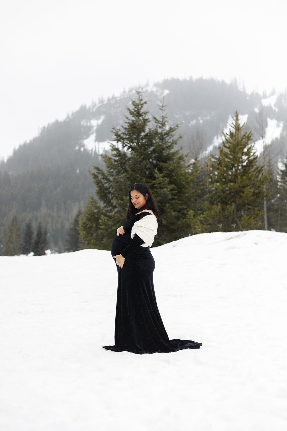 A person in a black coat stands in deep snow with evergreen trees and misty mountains in the background.