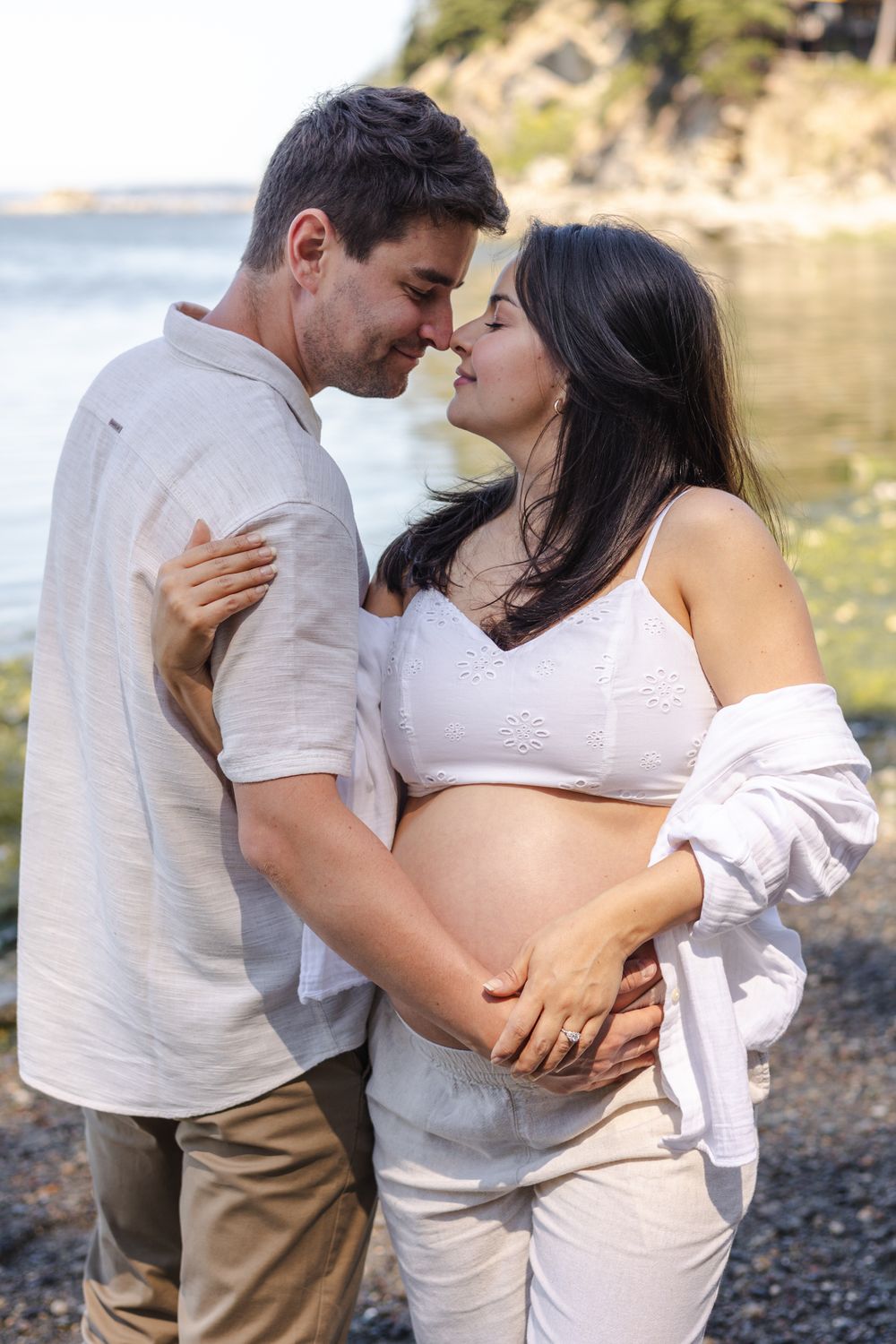 A couple in light colored clothing share romantic moments by a rocky shoreline during a photo session.