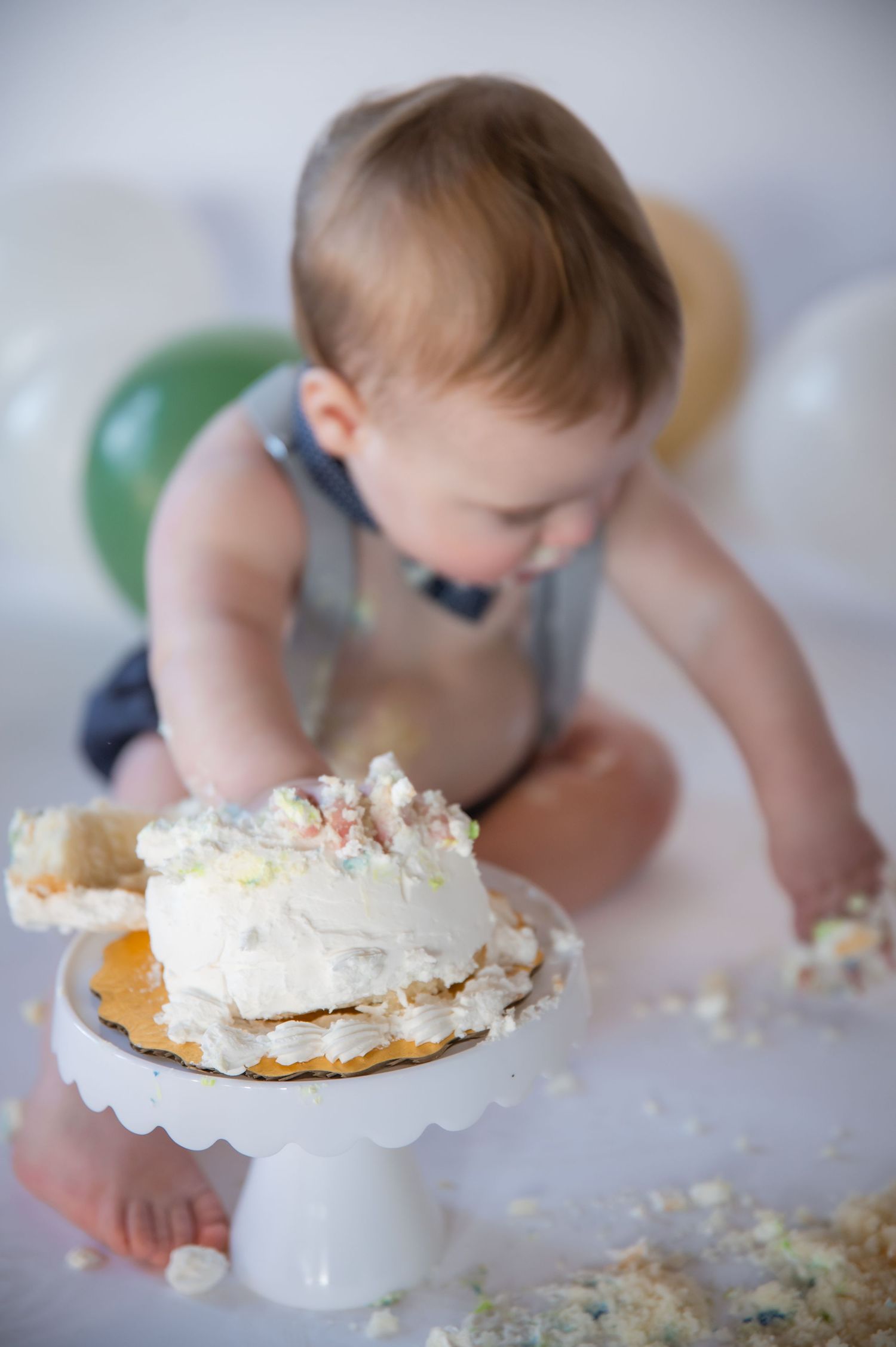 A baby in overalls curiously explores a white frosted cake placed on a pedestal cake stand during a cake smash photo session.