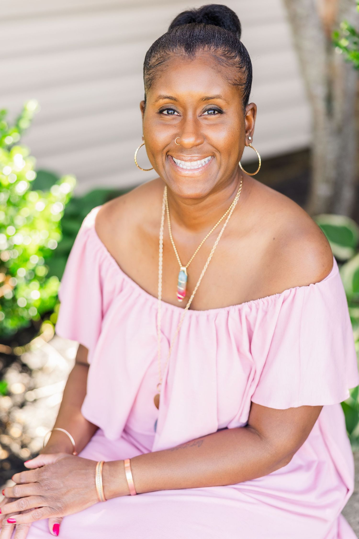 Smiling person in pink off-shoulder dress wearing layered necklaces and hoop earrings sitting outdoors.