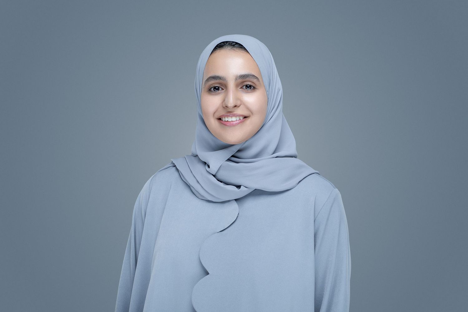A person in a light gray hijab and matching dress stands against a gray background with a warm expression.