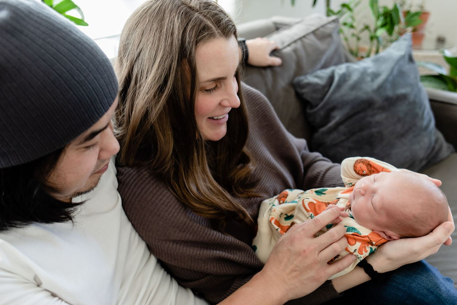 Caring adults sitting on couch bottle feeding a swaddled newborn baby while smiling.