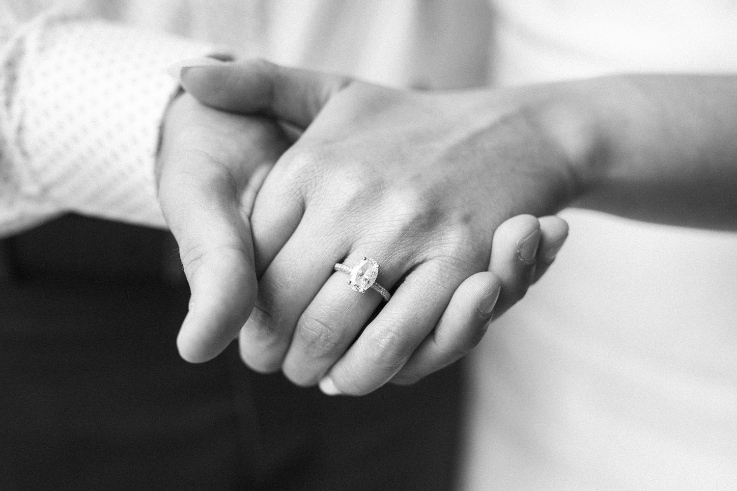 Black and white close-up of clasped hands showing engagement ring on finger.