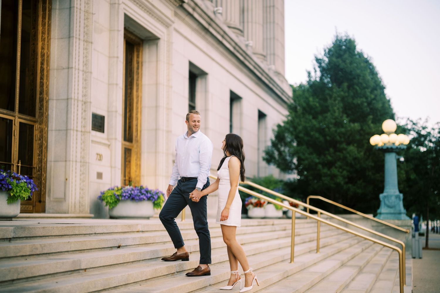 People holding hands on stone steps of a grand building entrance with ornate railing and lamp post in background.