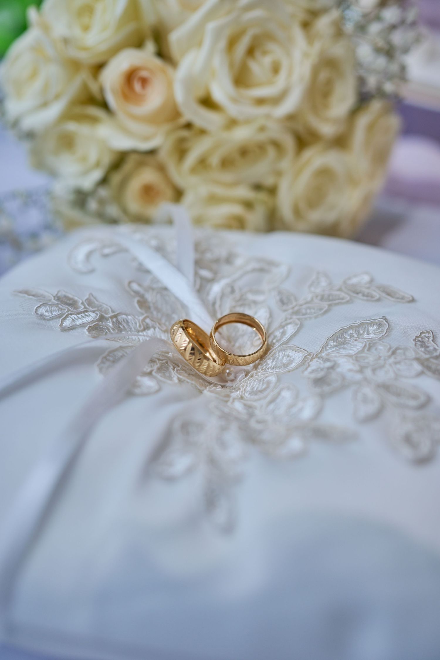 Gold wedding rings rest on delicate white lace fabric with cream roses in soft focus background.
