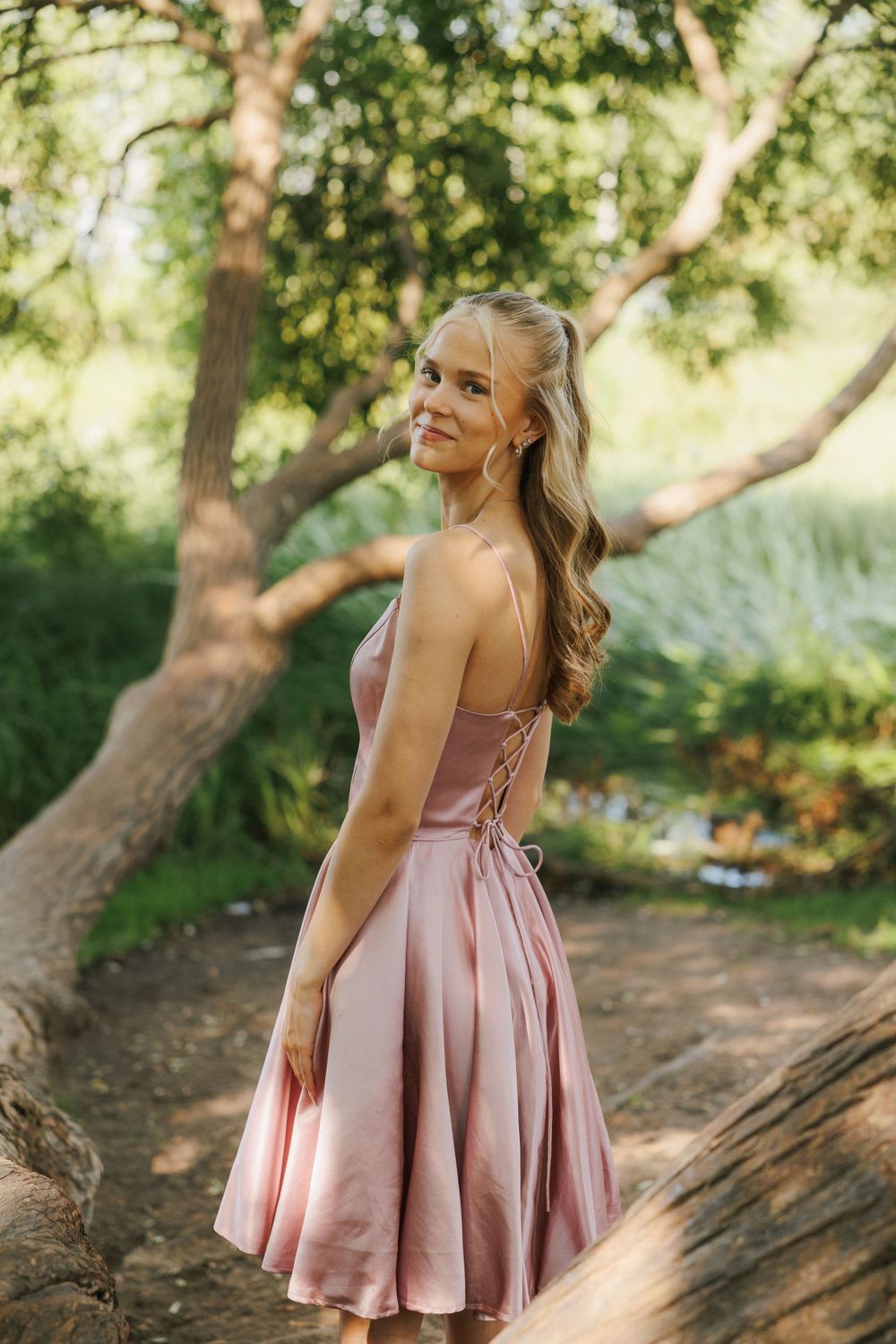 Person in a pink satin dress with lace-up back poses outdoors in a natural woodland setting.