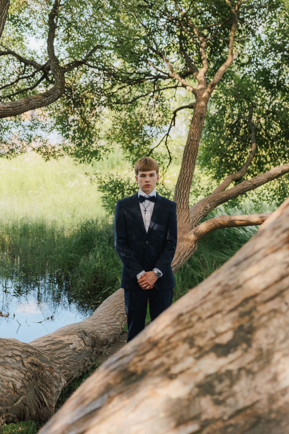 A person in a formal black tuxedo poses next to a large tree trunk by a peaceful pond on a sunny day.
