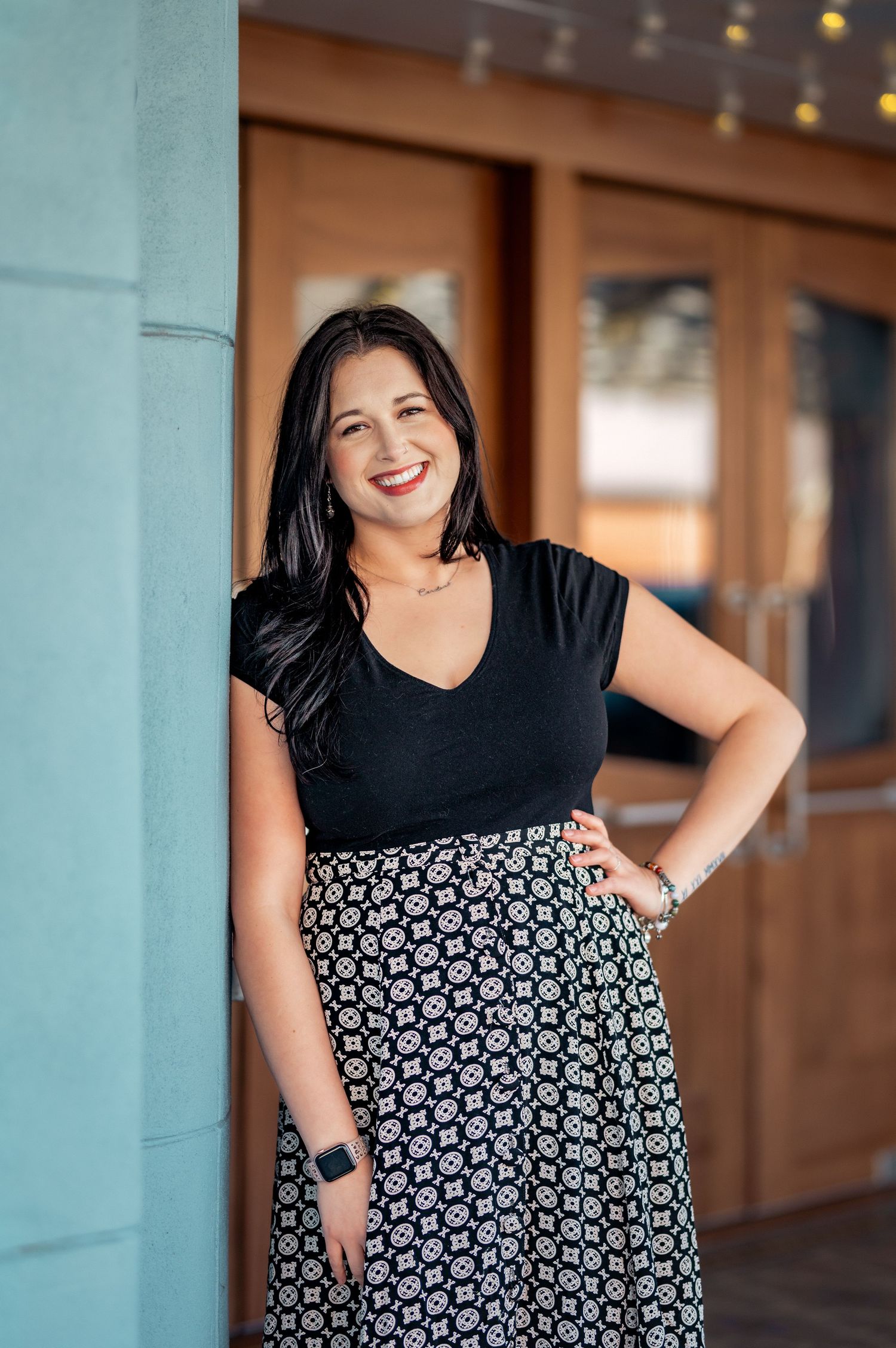 Someone wearing a black v-neck top and patterned skirt poses outside a building with string lights in background.