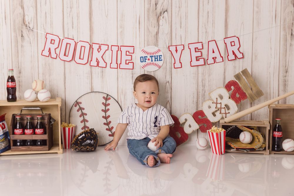 Vintage baseball themed baby photo setup with Rookie of the Year banner, bats, gloves and Coca-Cola decor against wooden wall.