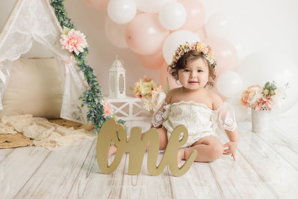 A smiling baby girl in a floral crown sits beside a golden 'one' sign at a first birthday photo shoot.