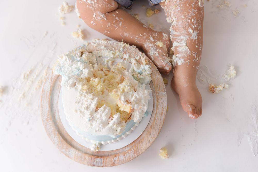 Small hands reach into a white cake smash setup with frosting and crumbs scattered on a wooden surface.