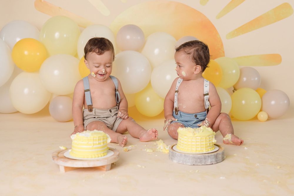 Two babies in diapers and suspenders sit with individual yellow birthday cakes against a pastel balloon backdrop.