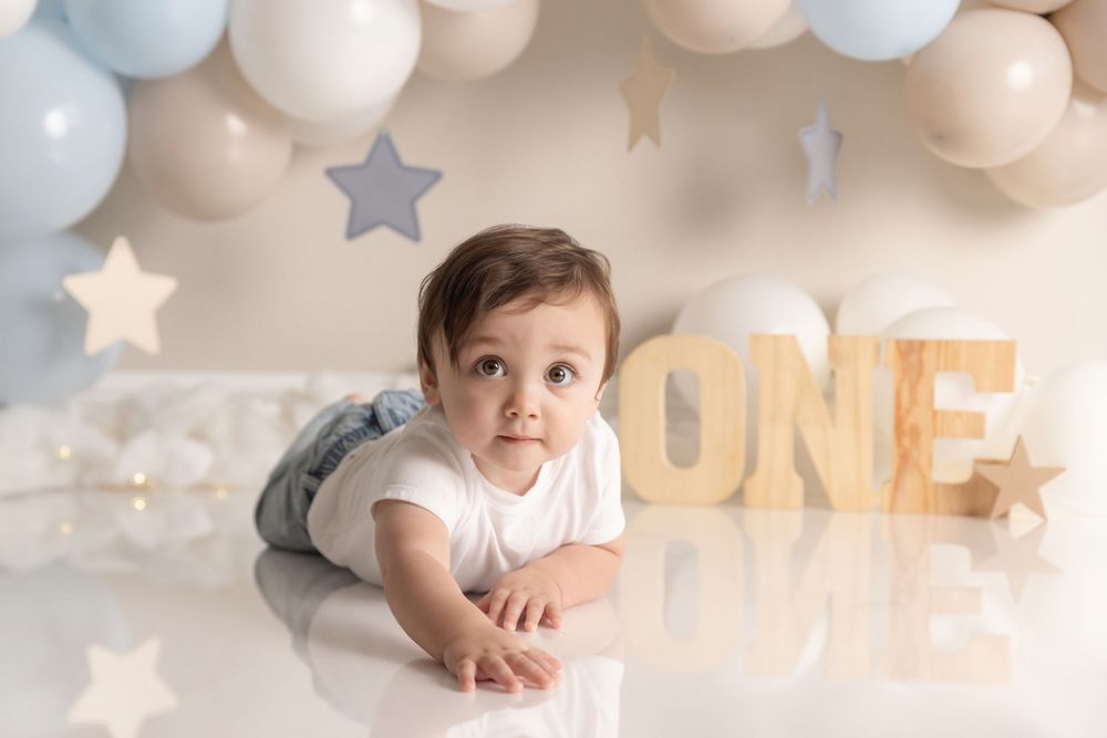 Baby in white outfit lies on floor with blue and beige balloon decorations and star accents for first birthday photo shoot.