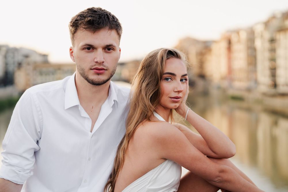 Narzeczeni siedzący razem na murku we Florencji, rozświetleni pierwszymi promieniami słońca, pełni szczęścia i czułości, Ponte Vecchio w tle