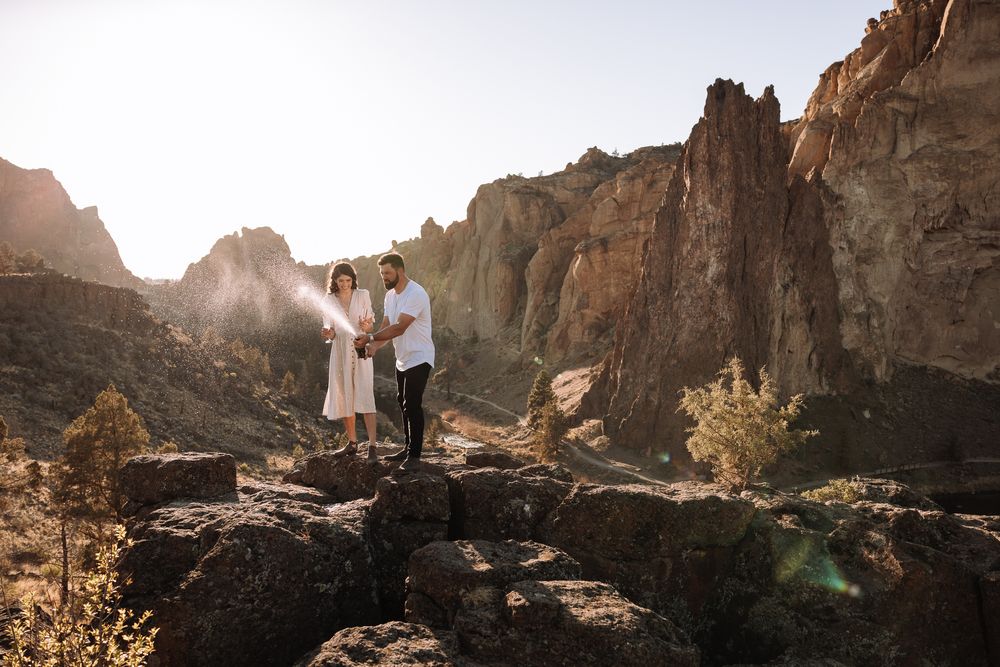 A couple stands together on rocky terrain with towering cliffs and sunlight in the background.
