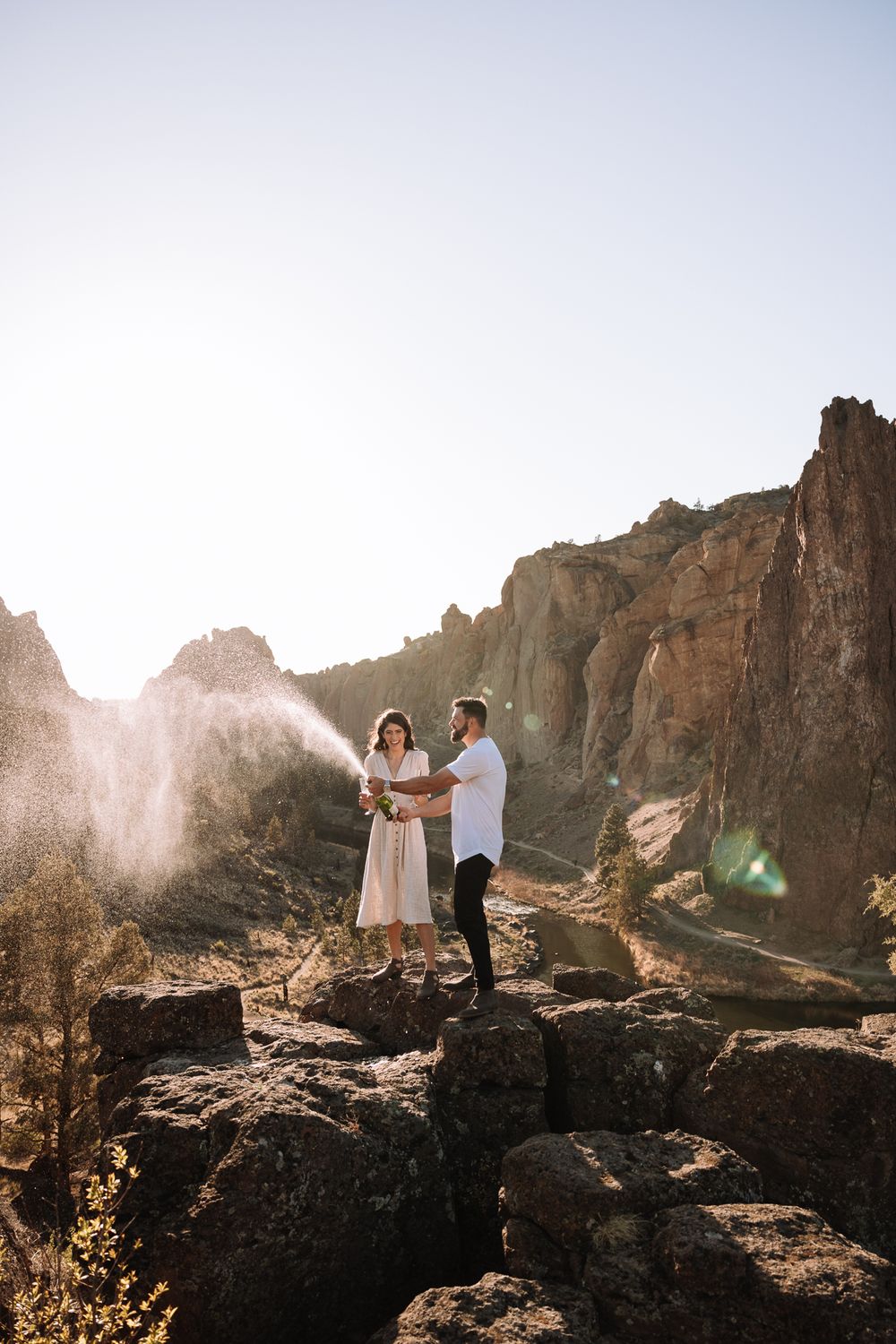 Two people embrace on a rocky cliff at Smith Rock State Park during golden hour with dramatic sunlight and mountain views.