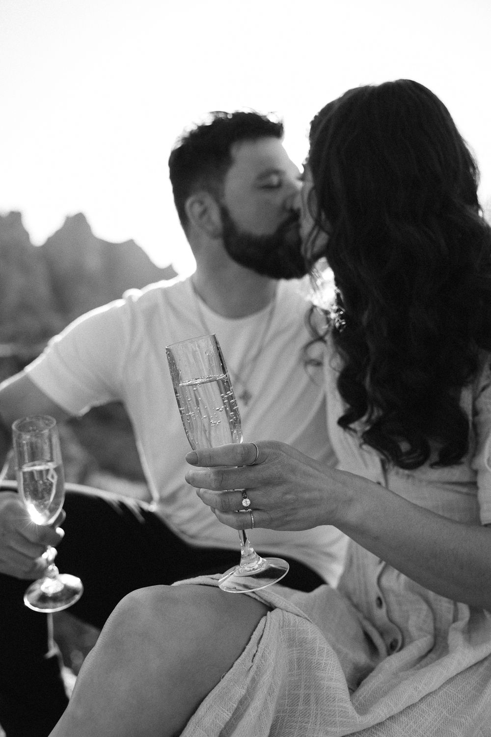 Black and white romantic moment with champagne glasses as couple shares intimate kiss during sunset outdoors.