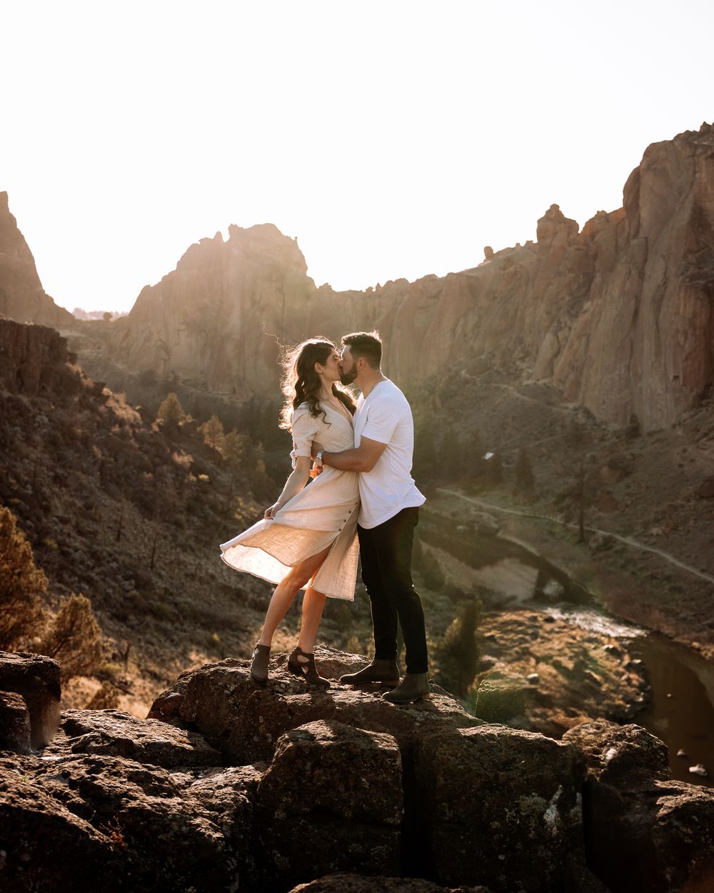 A couple embraces on a rocky overlook with dramatic canyon walls and golden sunlight in the background.