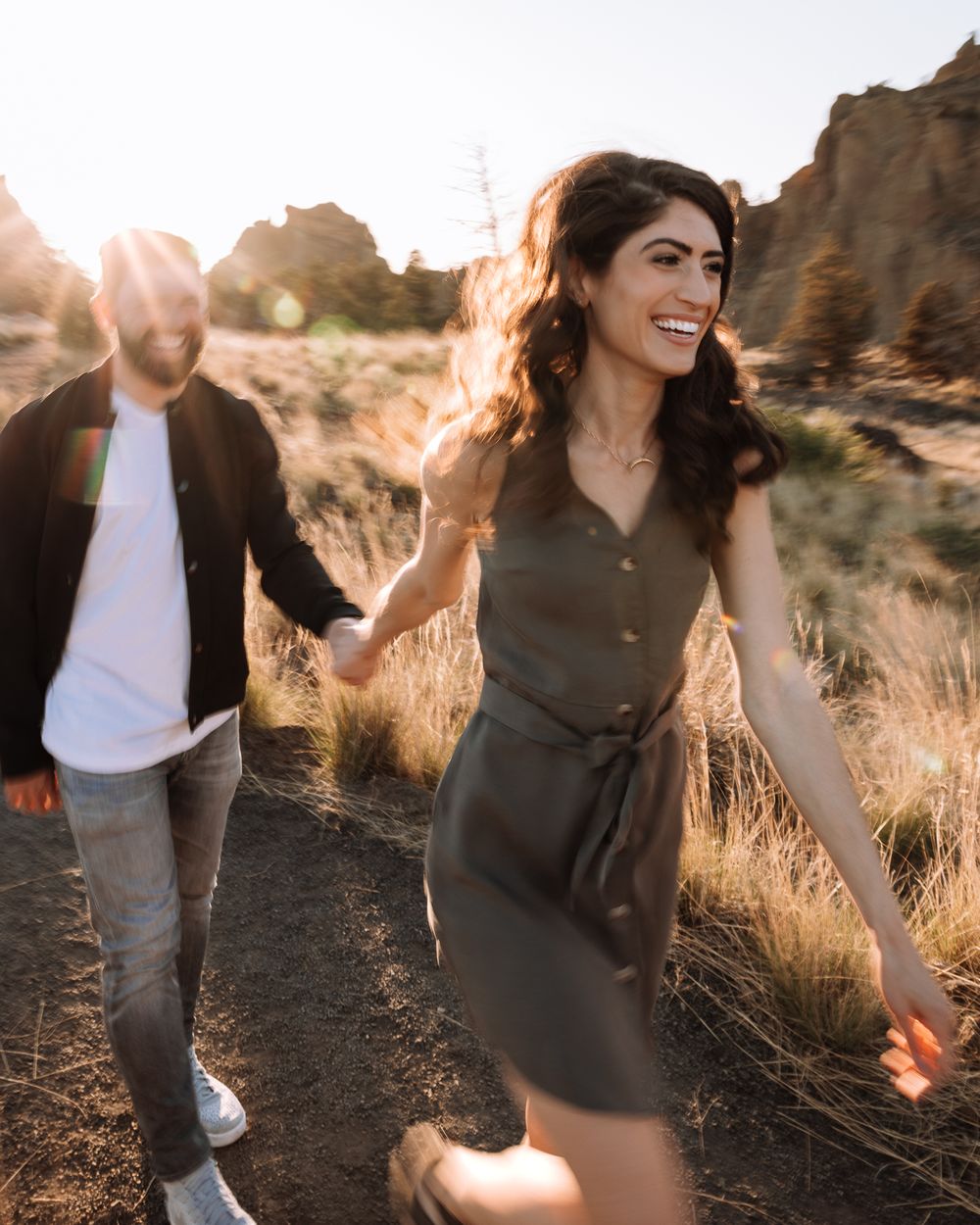 Two people holding hands and walking on desert trail at golden hour with sunflare and rock formations in background.