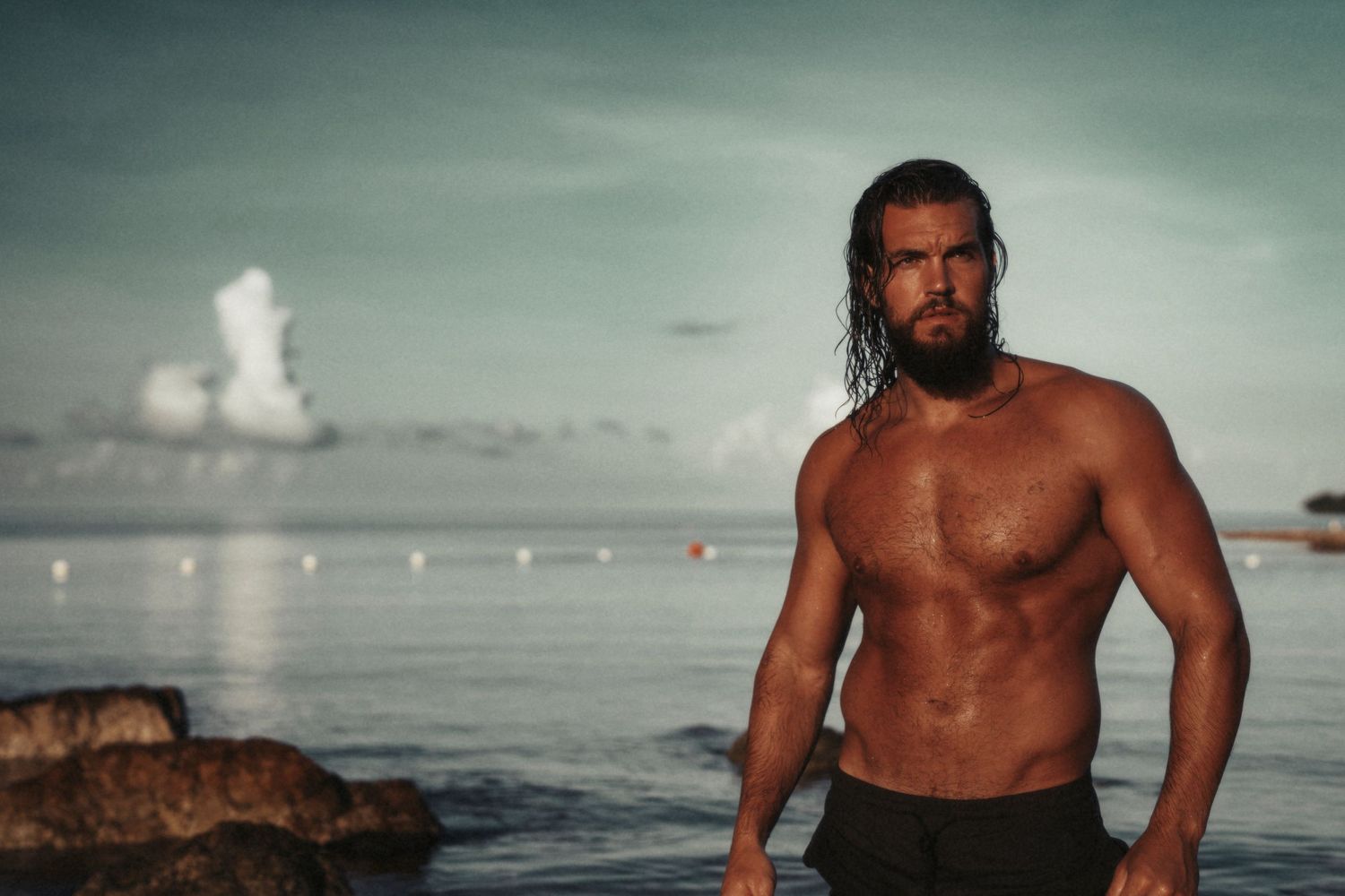 Athletic figure standing shirtless on rocky beach against moody ocean backdrop at sunset.