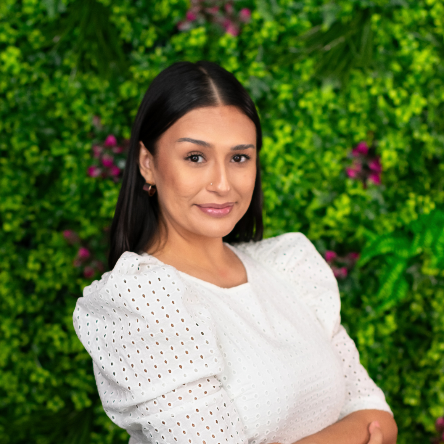 Professional portrait of a woman in a white eyelet blouse smiling warmly against a green garden backdrop.