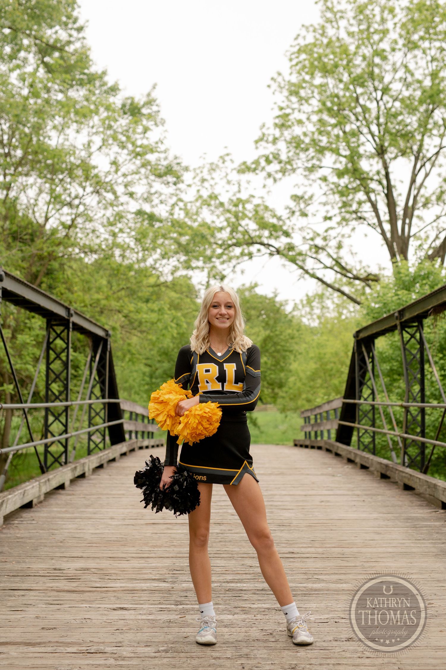 Cheerleader poses with black and yellow pom poms on a wooden bridge surrounded by green trees.