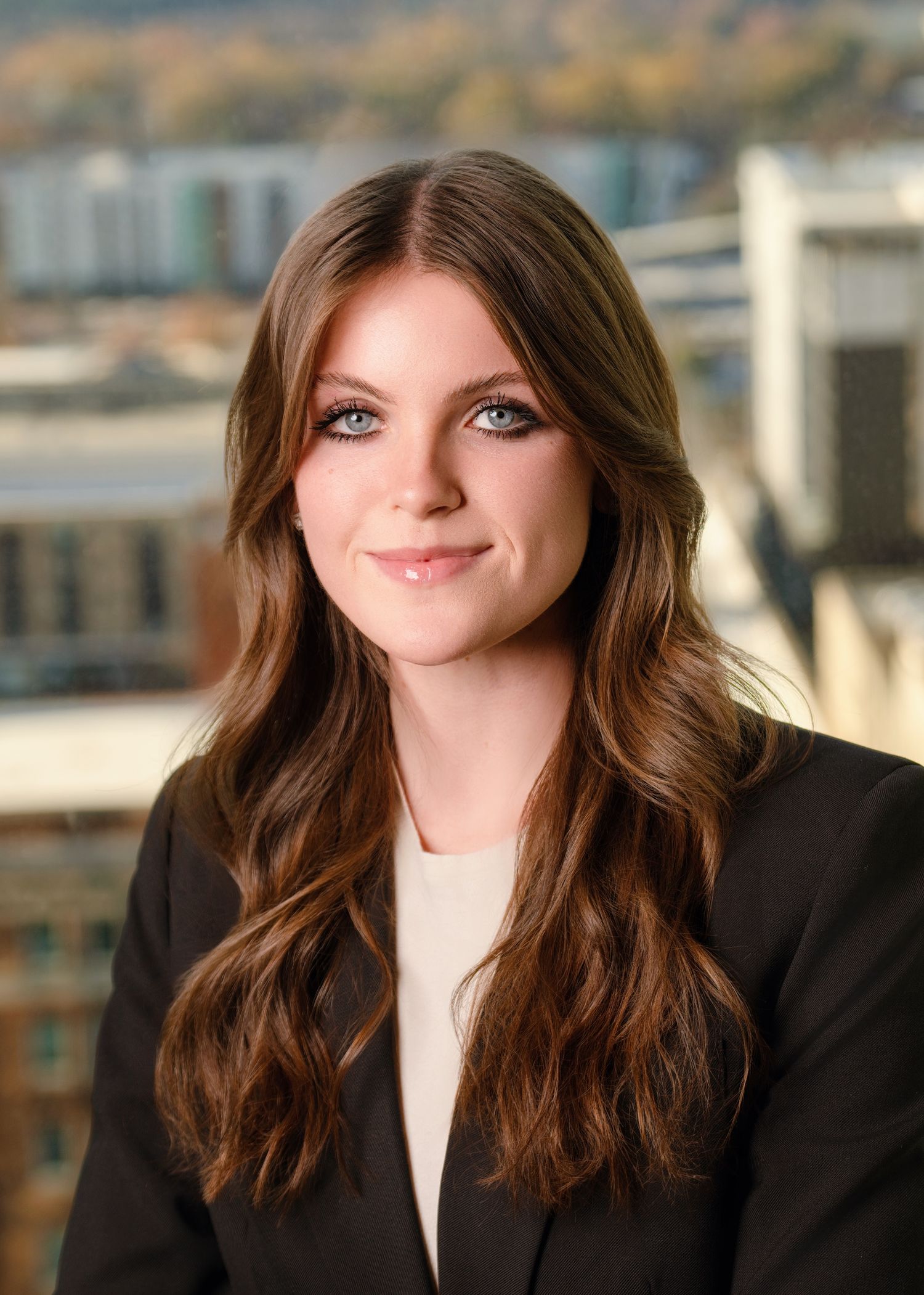 Professional headshot of a woman in a black blazer and cream top against a blurred cityscape background.