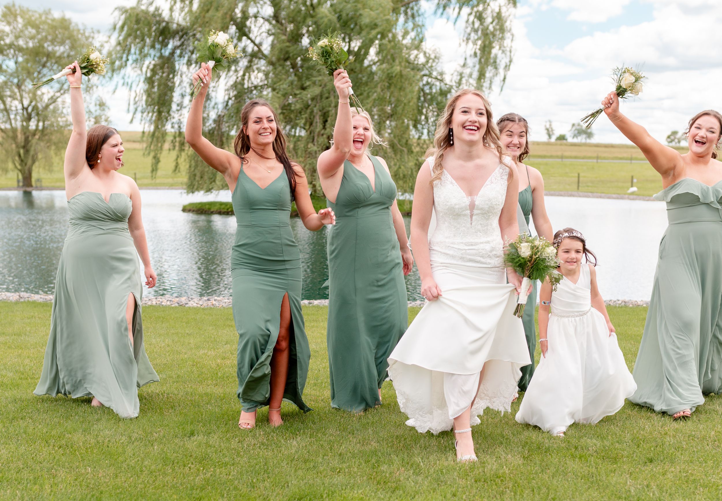 Wedding party in sage green bridesmaid dresses celebrating joyfully on grass near a pond.