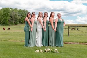 Wedding party wearing blue-green dresses stands together in a grassy field with horses grazing in the background.