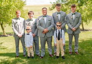 A group wearing gray suits and bow ties poses together on a sunny lawn during a wedding.