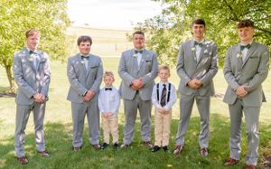 A group wearing matching light gray suits and bow ties pose together on grass during a wedding.