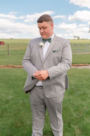A groom in a light grey suit adjusts his bowtie outdoors at a farm venue.