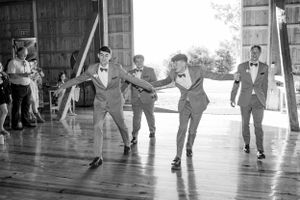 Black and white photo of groomsmen dancing energetically inside rustic barn reception venue.