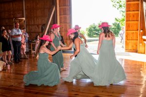 Wedding party members dancing together in matching sage green dresses and pink cowboy hats at a rustic barn venue.