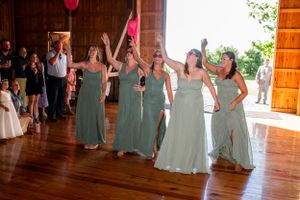 Bridesmaids in sage green dresses dancing and celebrating at a rustic indoor wedding reception.