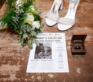 Wedding details featuring sparkly shoes, floral bouquet, announcement paper and ring box on wooden surface.
