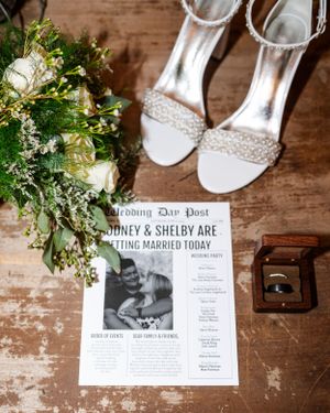 Wedding details flatlay with white heels, greenery bouquet and newspaper-style wedding announcement.